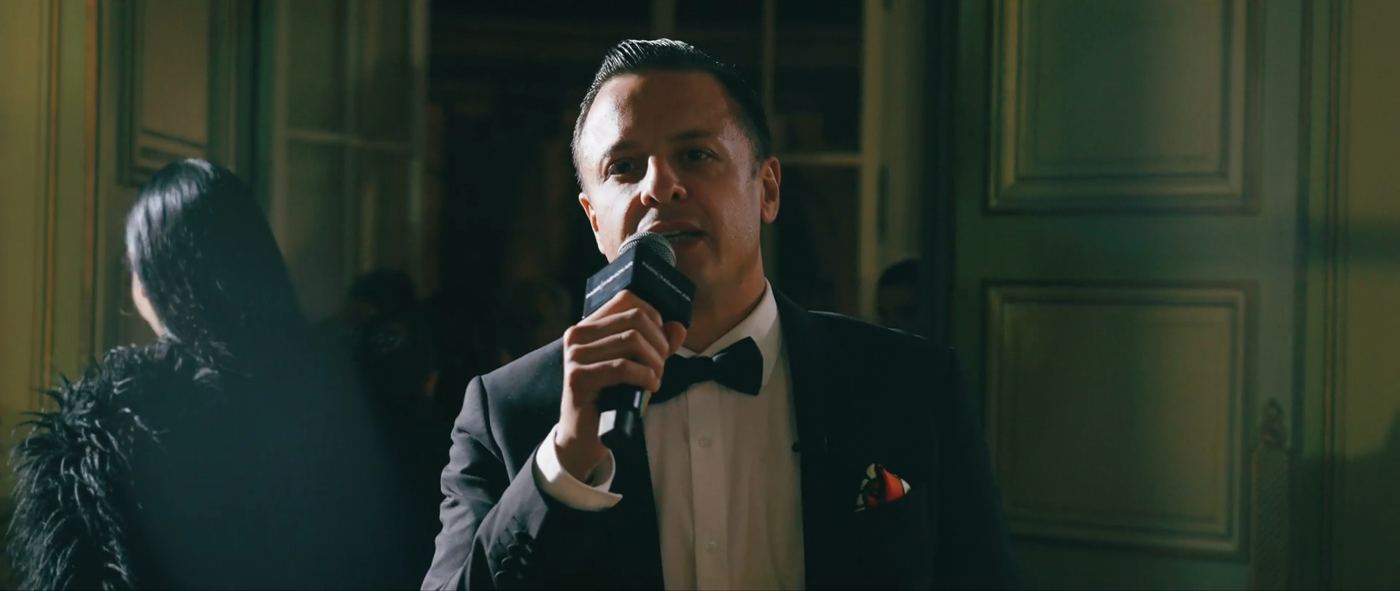Naïm Terrache in a dinner jacket speaking into a handheld microphone in a panelled room