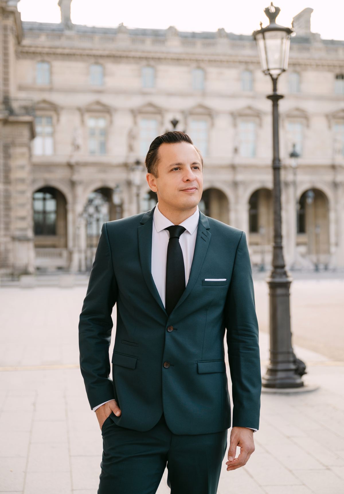 Portrait of Naïm Terrache in a dark suit, hands in pockets, in the courtyard of the Louvre