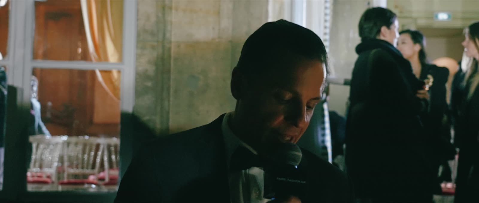 Naïm Terrache speaking into a microphone in a dim room, guests behind him