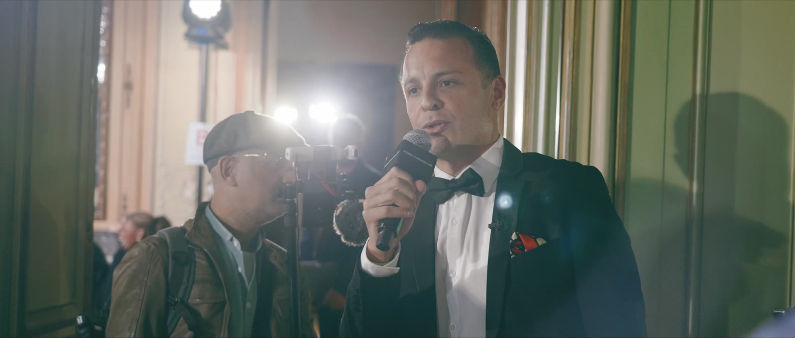 Naïm Terrache speaking into a microphone at a fashion show, photographers and lights behind him