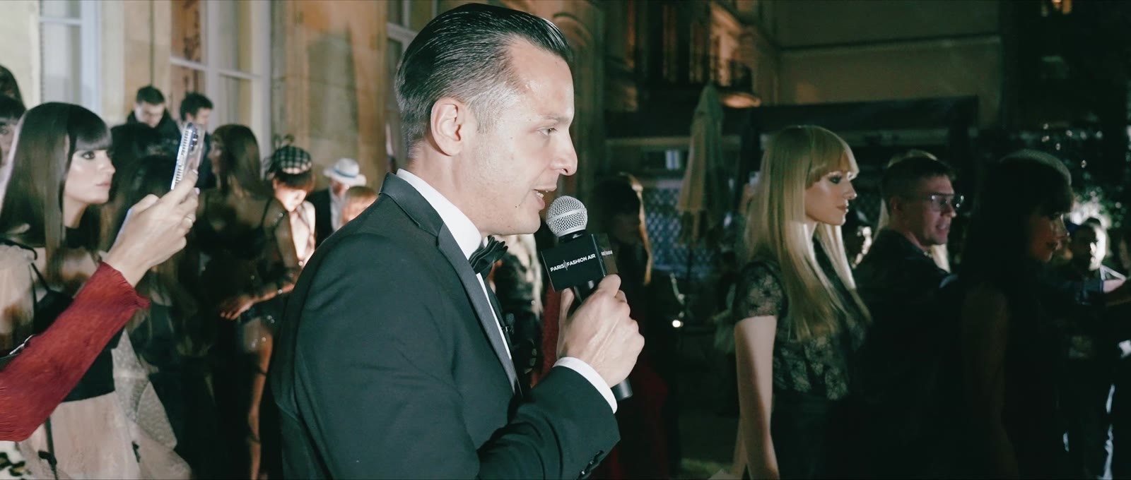 Naïm Terrache hosting a fashion show, microphone in hand, guests filming with their phones